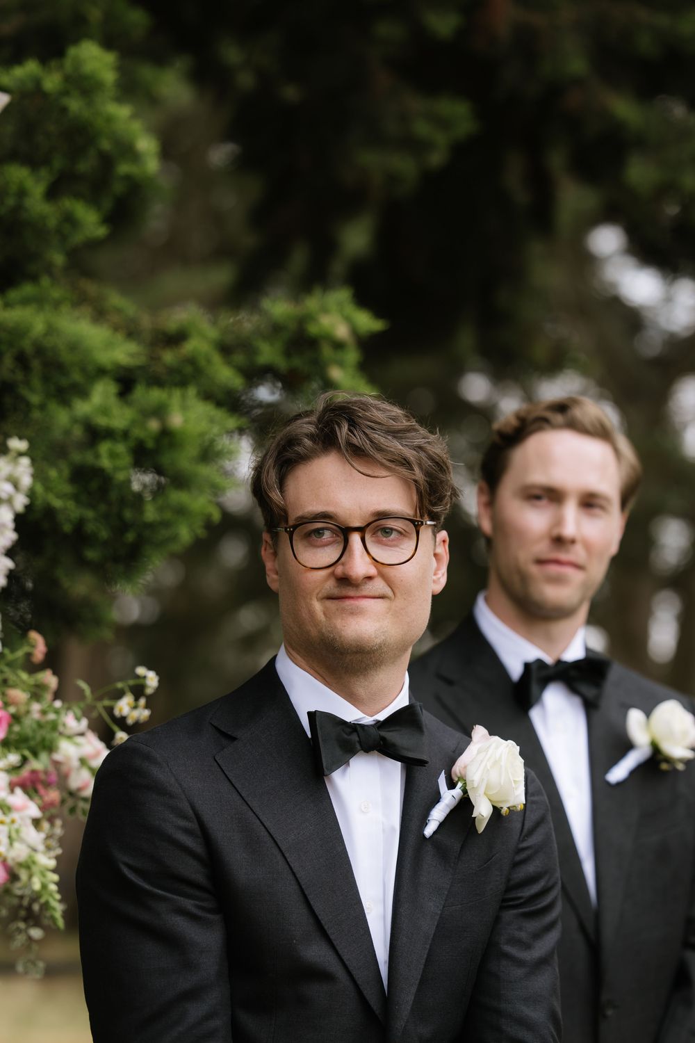 Two groomsmen in black tuxedos with bow ties and boutonnieres stand outdoors at a wedding ceremony.