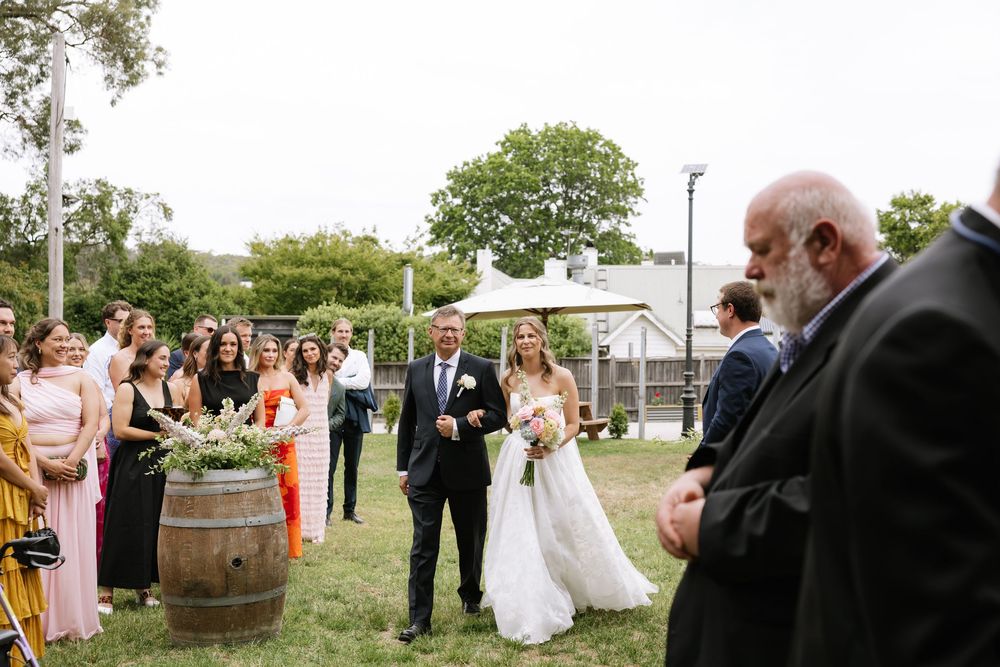 Bride walks down outdoor aisle with father at rustic wedding ceremony, guests watching beside wine barrel decorations.
