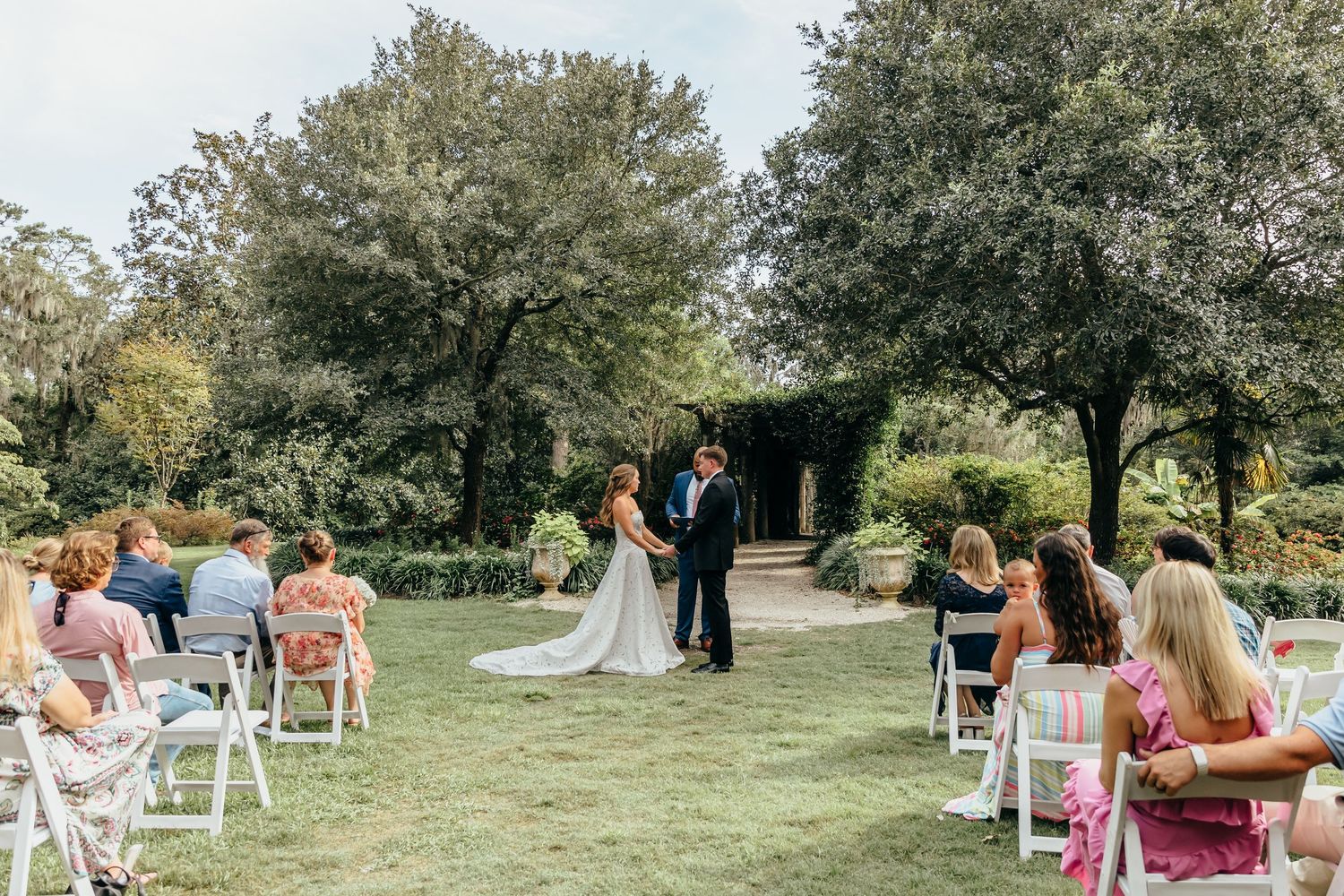 Outdoor garden wedding ceremony with bride and groom at the altar, guests seated on white chairs on a lush green lawn.