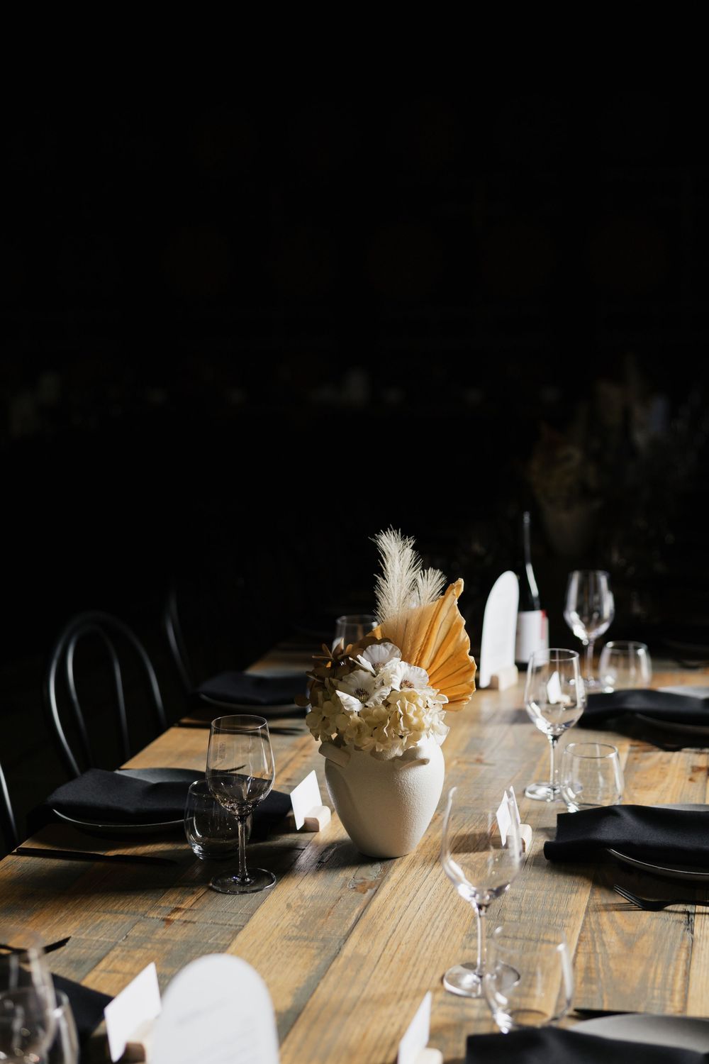 Elegant rustic wooden table set with black napkins, crystal glasses, and a white vase centerpiece with dried florals.