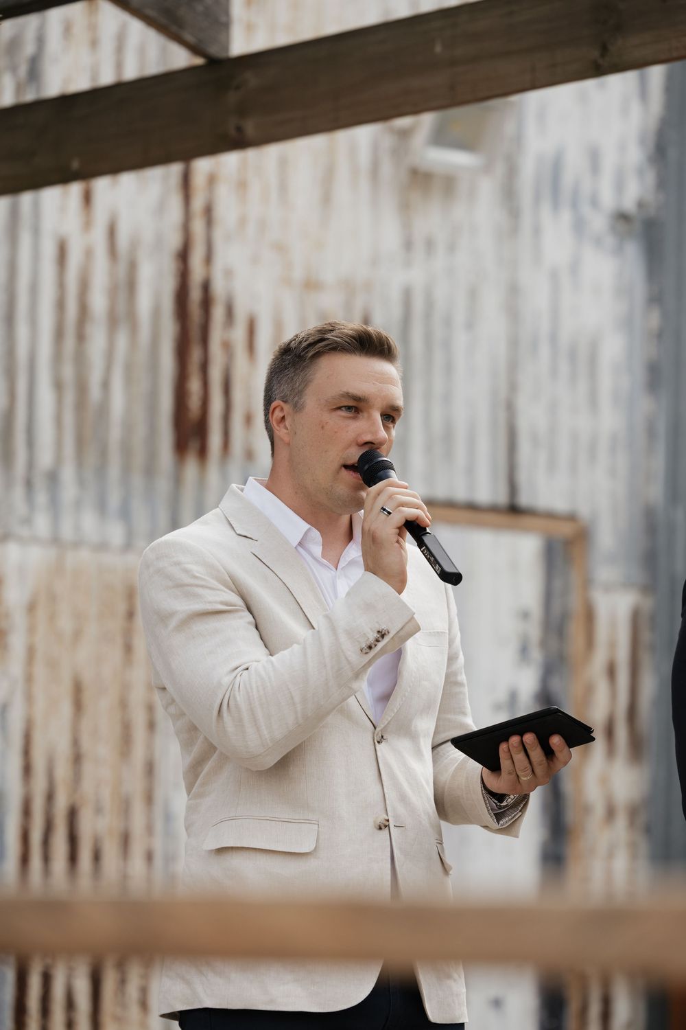 Man in white blazer speaking into microphone outdoors, holding tablet at rustic event venue.