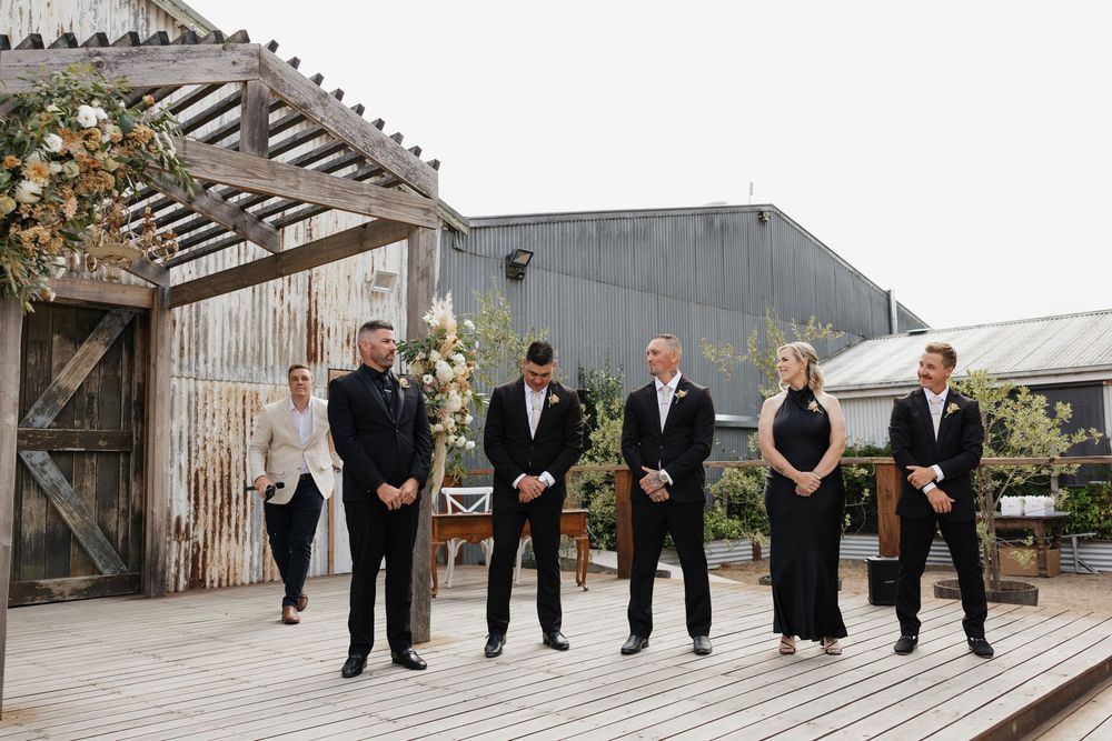 Wedding party in black attire standing on rustic wooden deck with floral arch backdrop at outdoor venue.