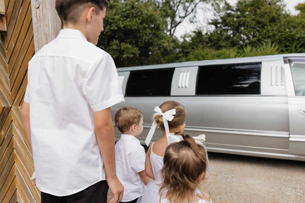 Three children in white outfits admiring a silver stretch limousine outdoors.