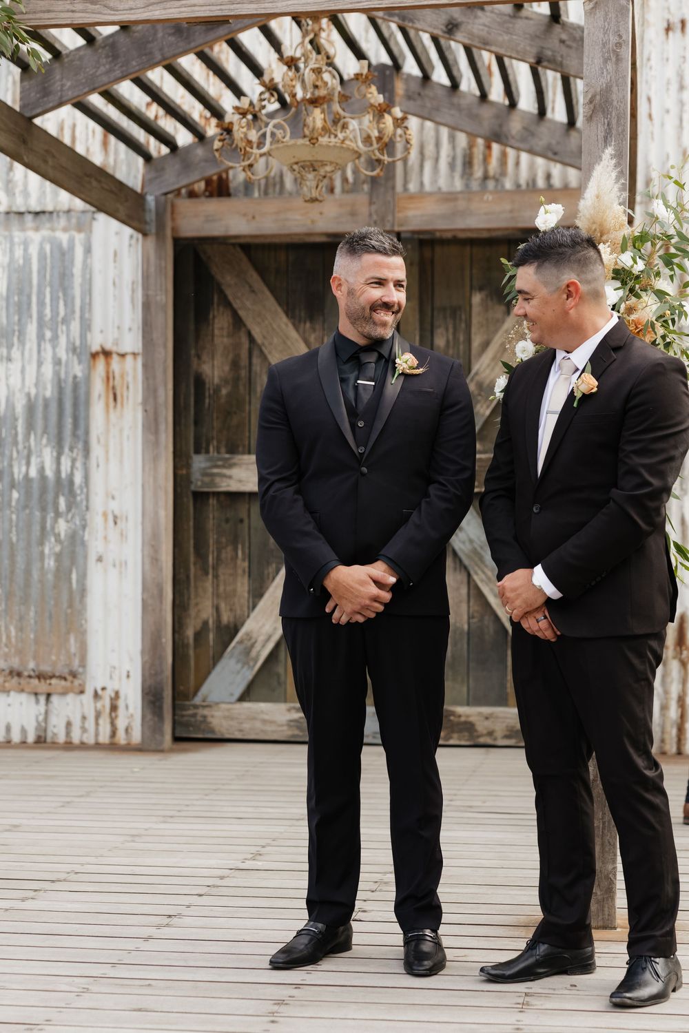 Two grooms in black tuxedos smile at each other during an outdoor wedding ceremony beside rustic barn doors.