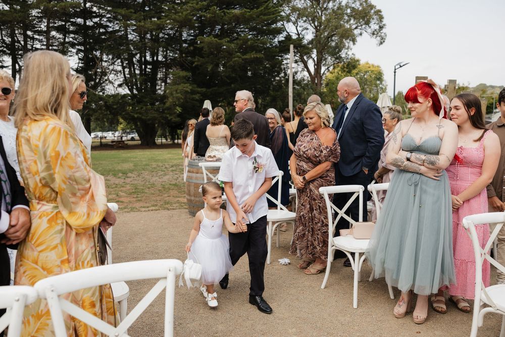 A young flower girl walks down the aisle with a groomsman at an outdoor wedding ceremony with seated guests.
