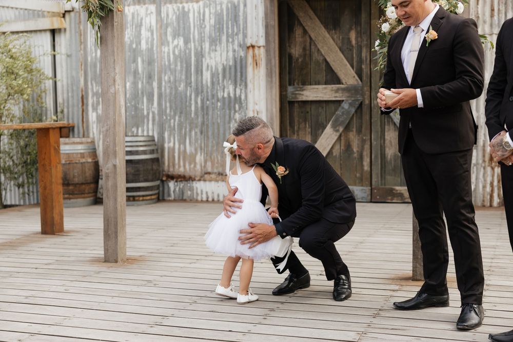 Man in black suit crouching to greet small flower girl in white dress at rustic outdoor wedding ceremony.