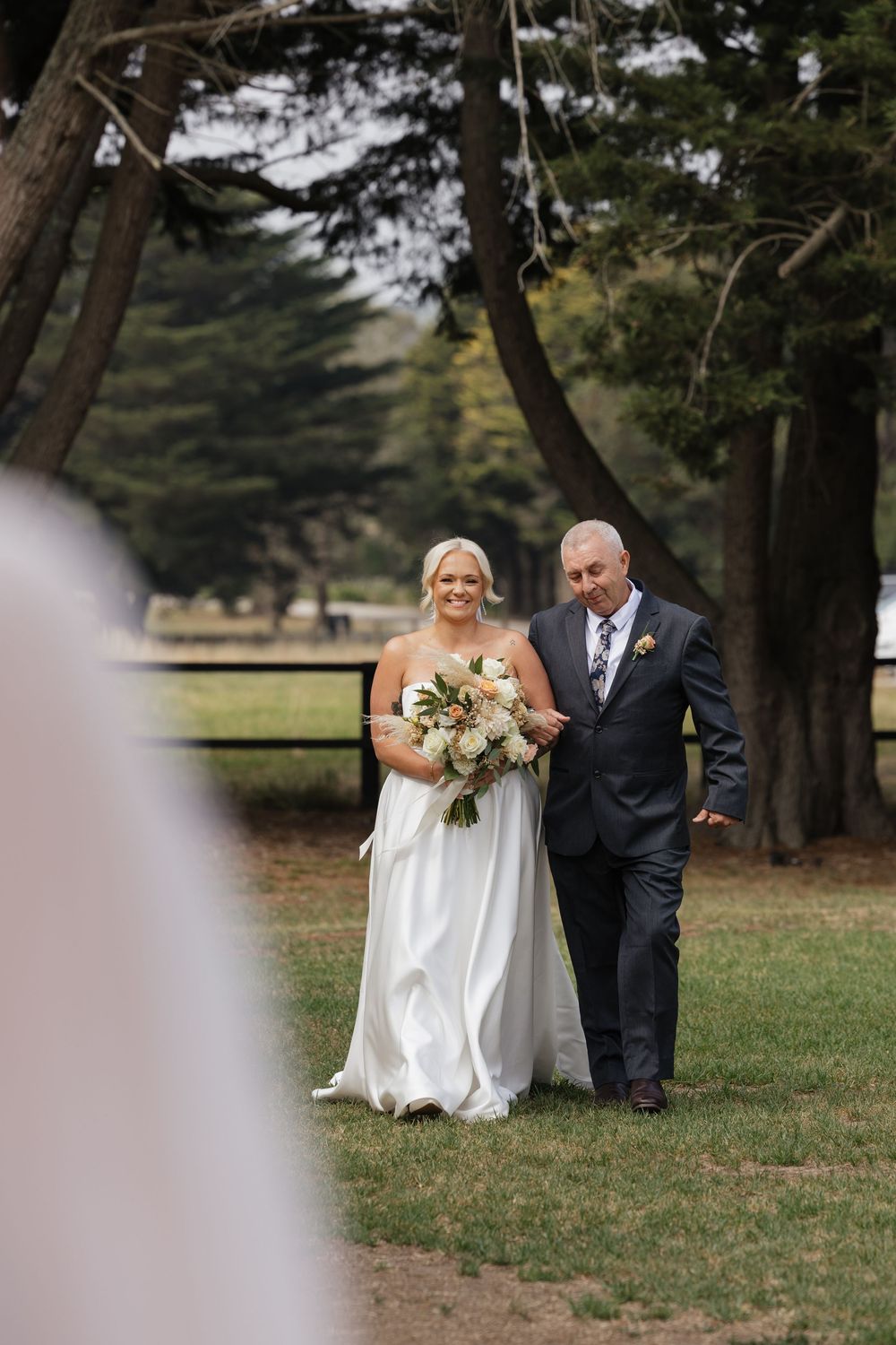 Bride in white gown holding bouquet walks down outdoor aisle with escort among tall trees at garden wedding.
