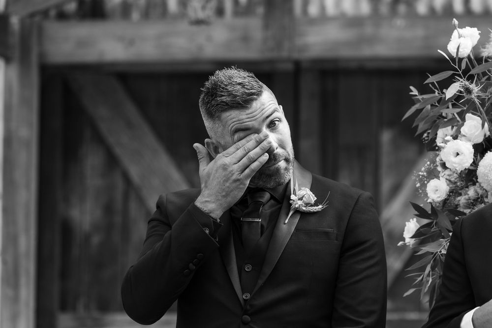 Black and white photo of emotional groom wiping tears at wedding altar with floral arrangement nearby.