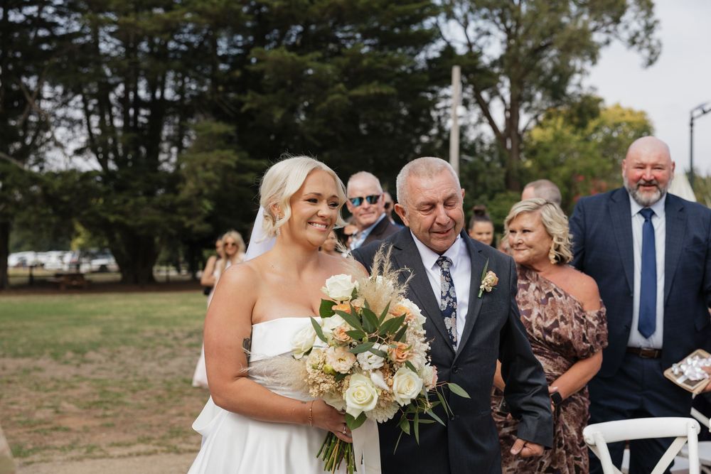Bride in white gown holding bouquet walks down outdoor aisle with older man at sunny wedding ceremony.