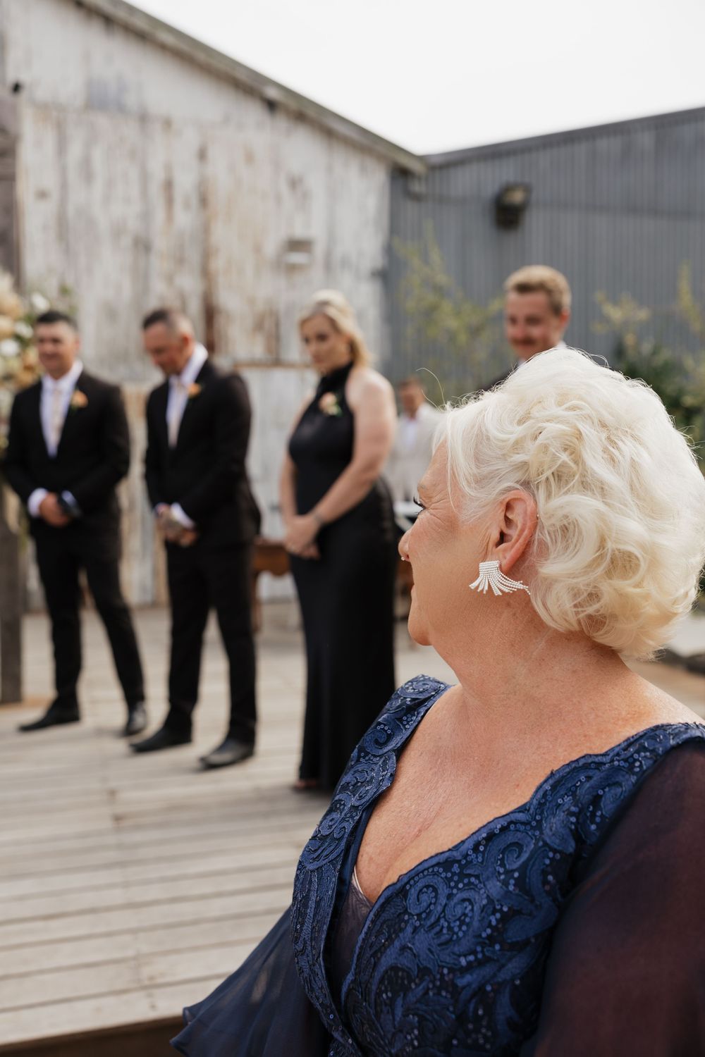 Elderly woman in blue lace dress watches wedding party in black attire standing on rustic outdoor venue deck.