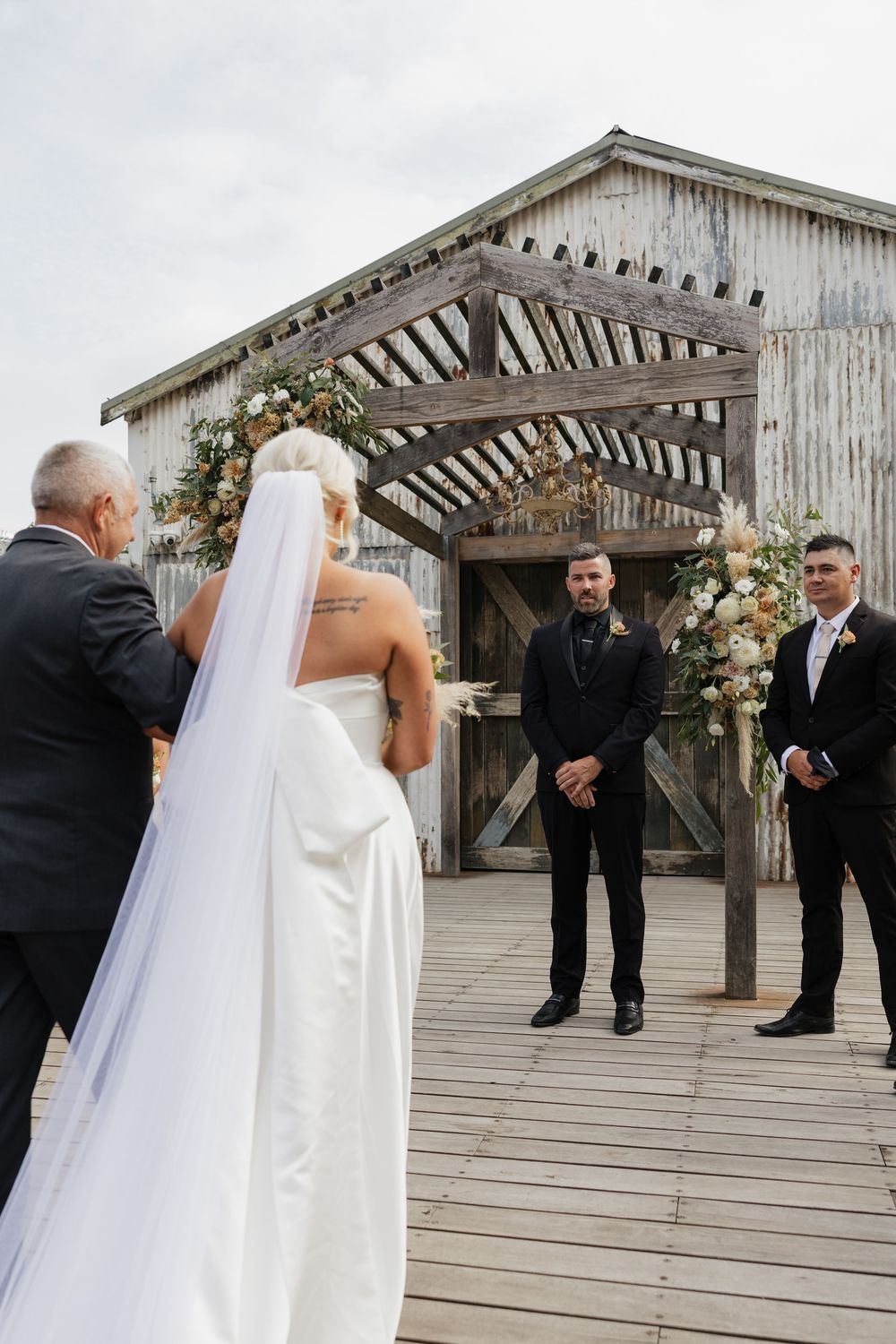 Bride in white gown and veil walks toward groom at rustic barn wedding ceremony with floral arch decorations.