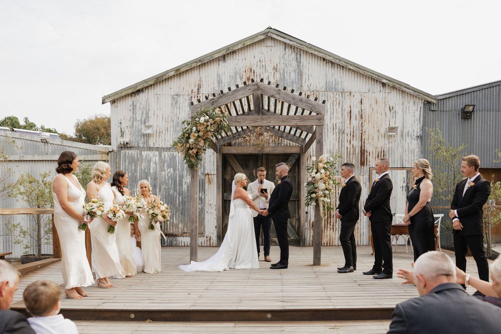 Outdoor wedding ceremony on a wooden deck in front of a rustic barn with bridal party and guests.