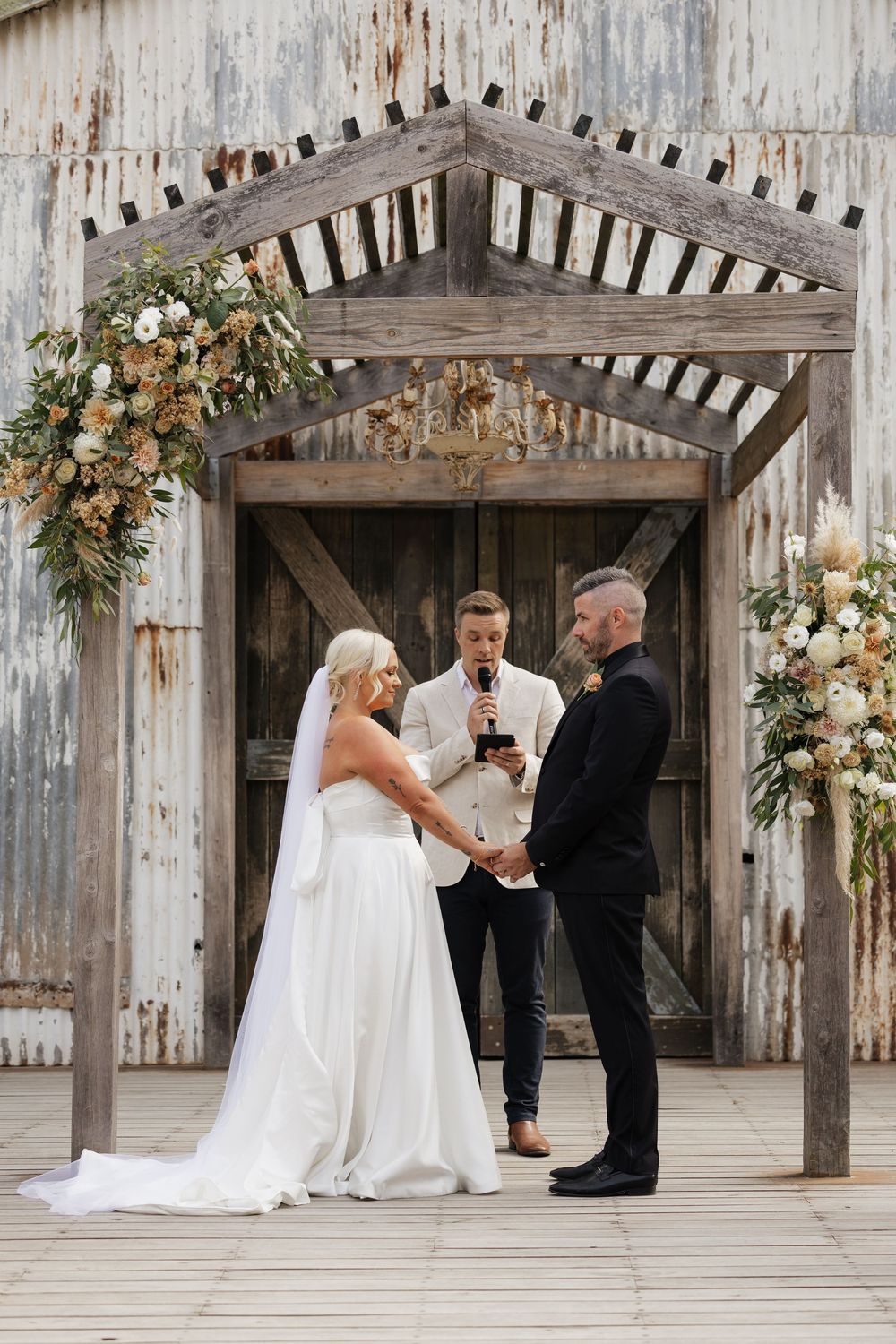 Bride and groom exchange vows before officiant at rustic barn wedding altar adorned with neutral floral arrangements.