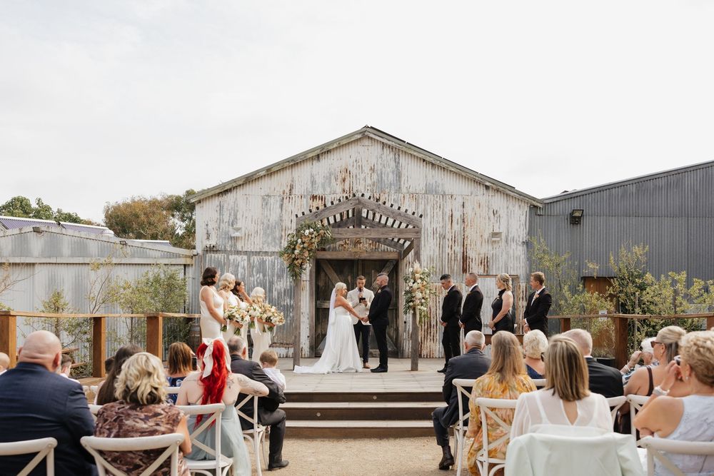 Outdoor wedding ceremony at a rustic barn venue with wedding party at the altar and guests seated in white chairs.