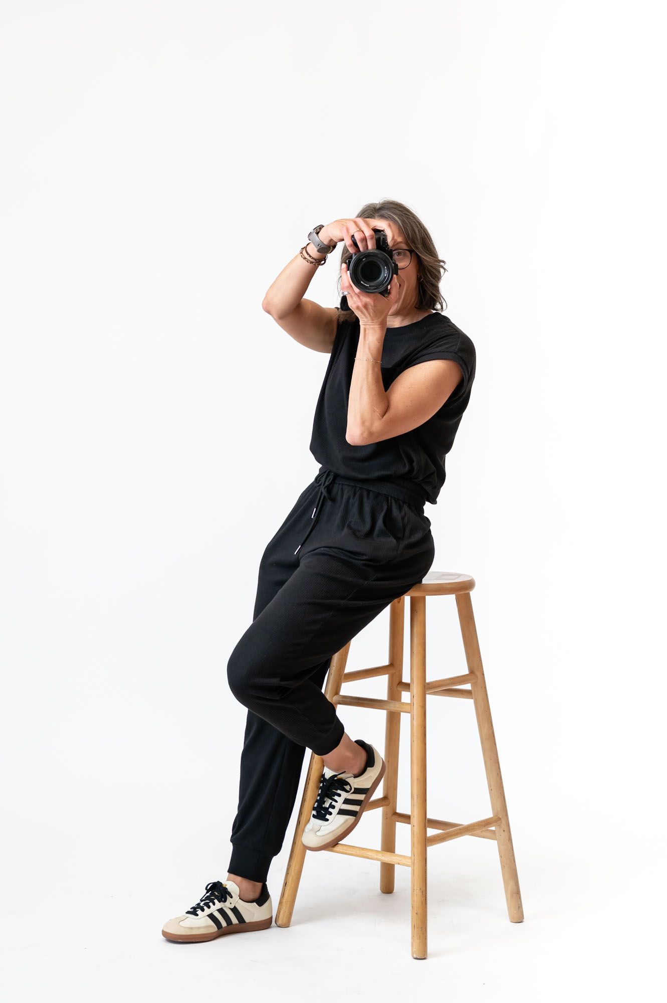 Photographer in black jumpsuit and Adidas sneakers sitting on wooden stool, holding DSLR camera up to face.