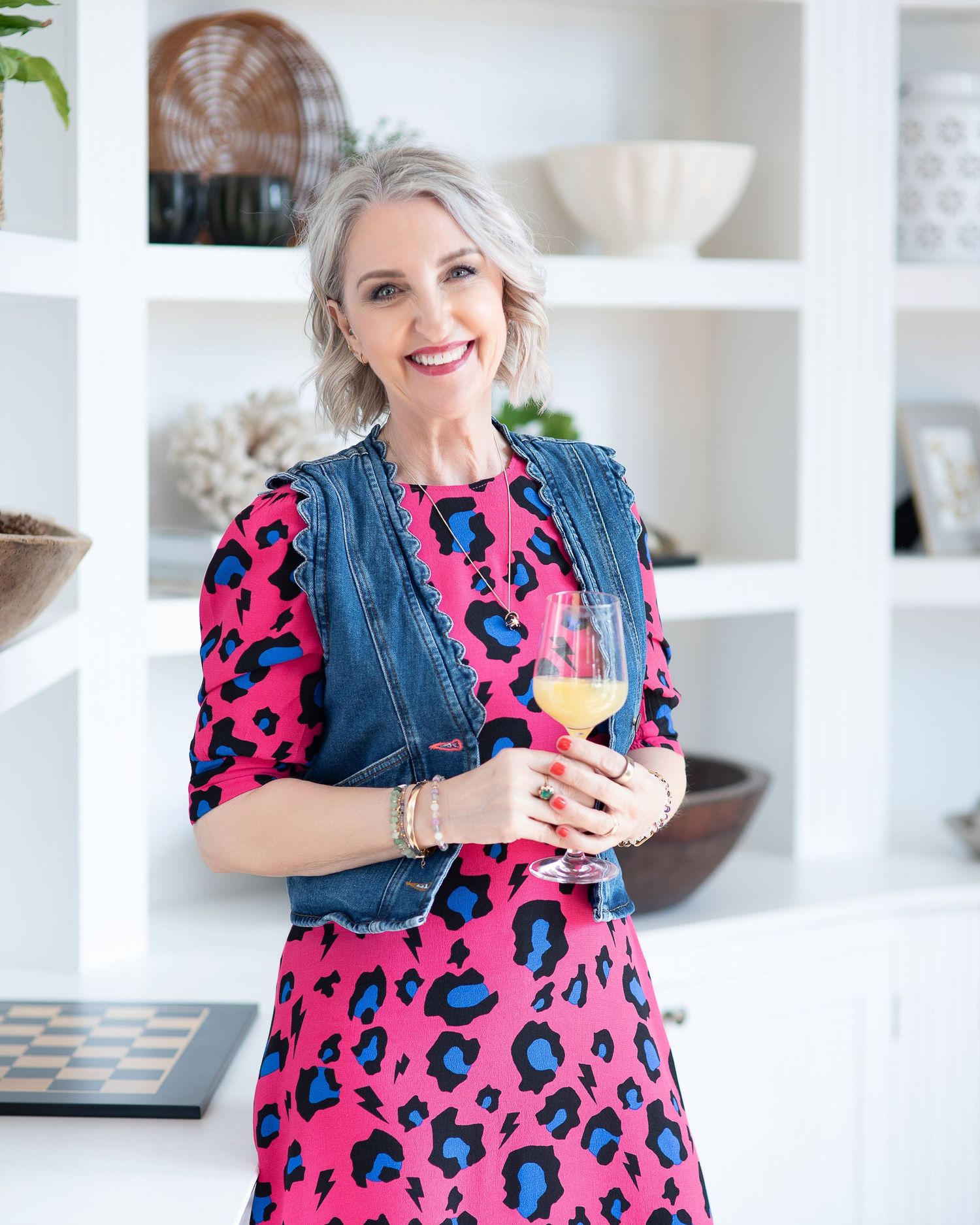 Woman in pink leopard print dress and denim vest holding white wine glass, smiling in bright white kitchen.