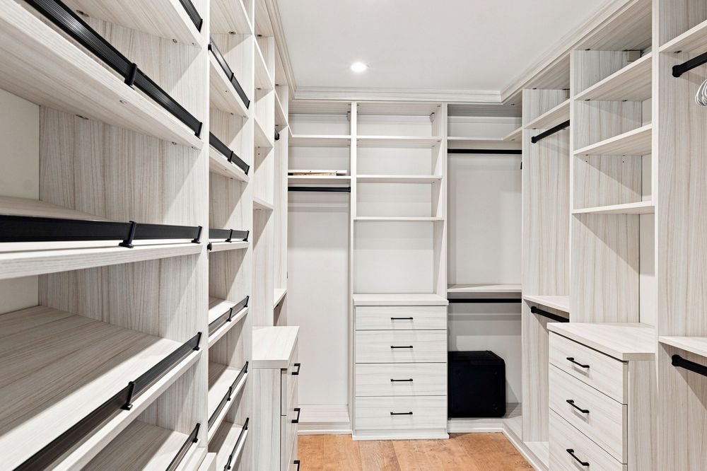 Spacious walk-in closet with white built-in shelving, hanging rods, drawers, and light hardwood floors.
