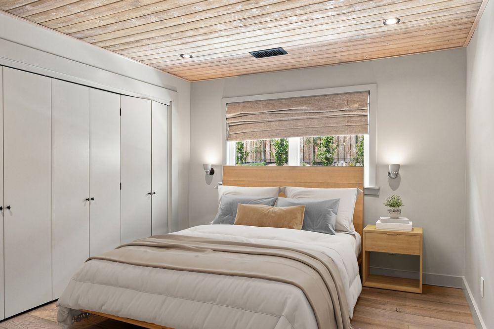 Cozy bedroom with wood plank ceiling, white built-in wardrobe, queen bed with neutral bedding, and bamboo roman shade window.