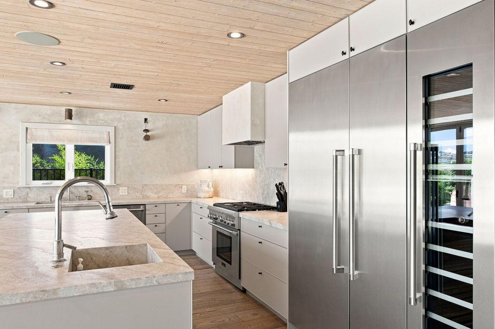 Modern kitchen with stainless steel appliances, white cabinets, marble countertops, and wood plank ceiling.