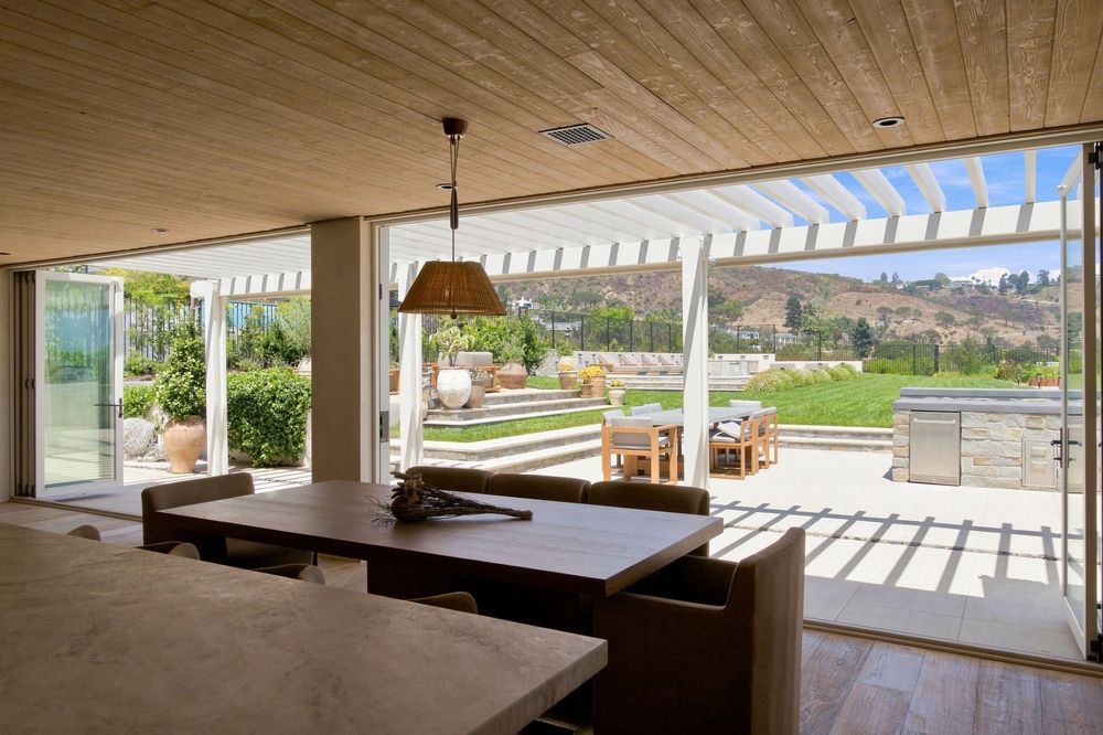Modern indoor-outdoor living space with sliding glass doors opening to a sunny patio with pergola and scenic hillside views.