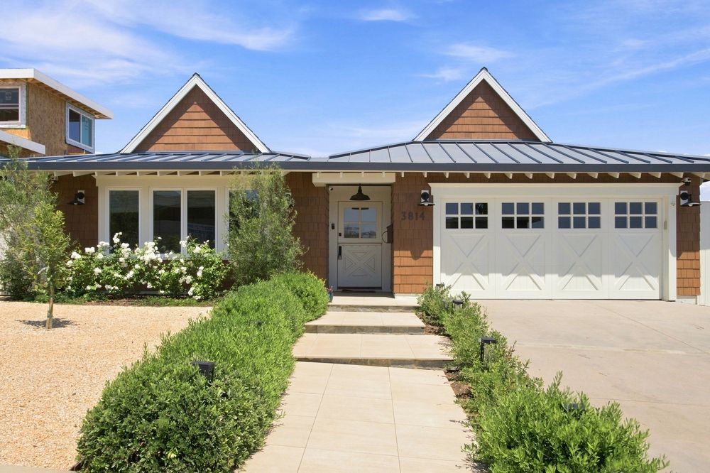 Modern ranch-style home with cedar siding, metal roof, white garage door, and lush landscaped front yard under blue sky.