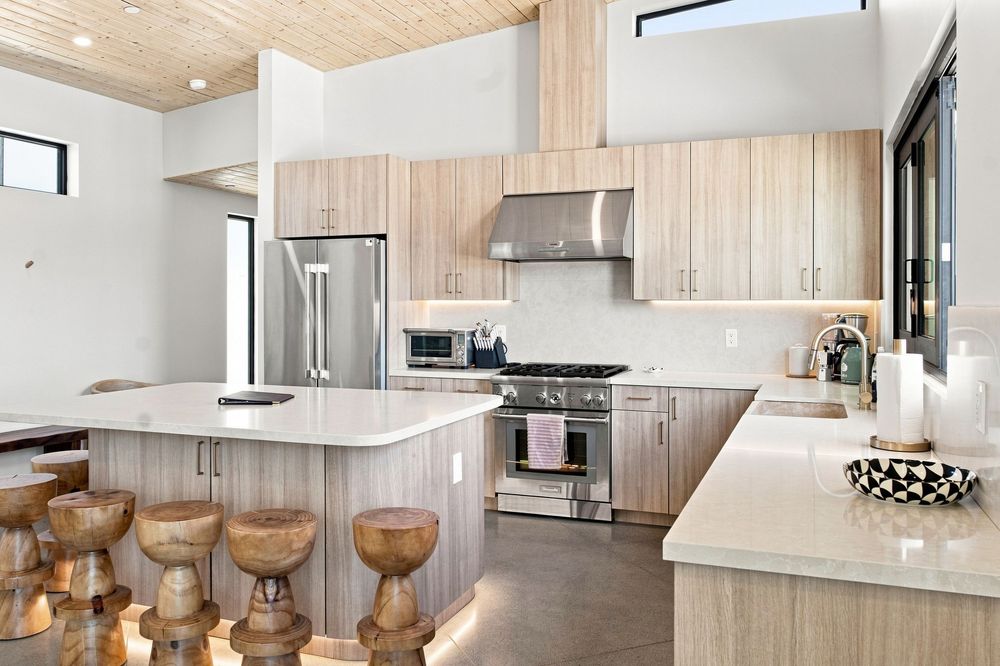 Modern kitchen with light wood cabinets, large island, wooden bar stools, stainless steel appliances, and white quartz countertops.