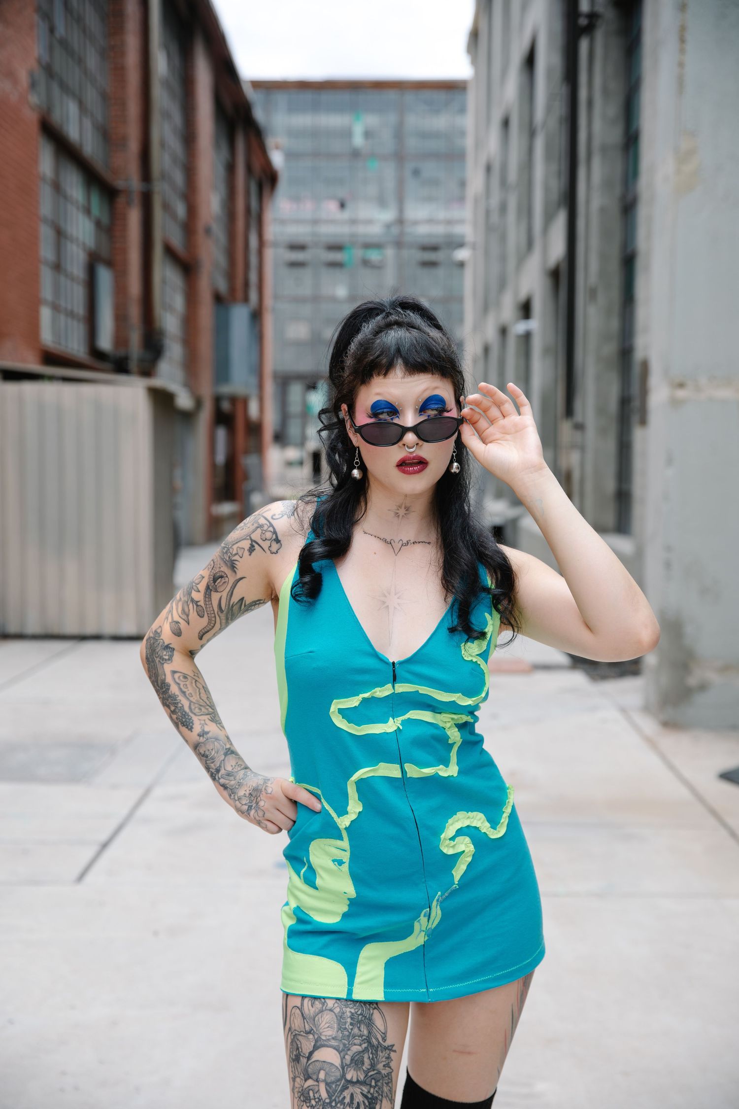 Tattooed woman in teal mini dress and blue sunglasses poses in urban alley setting.