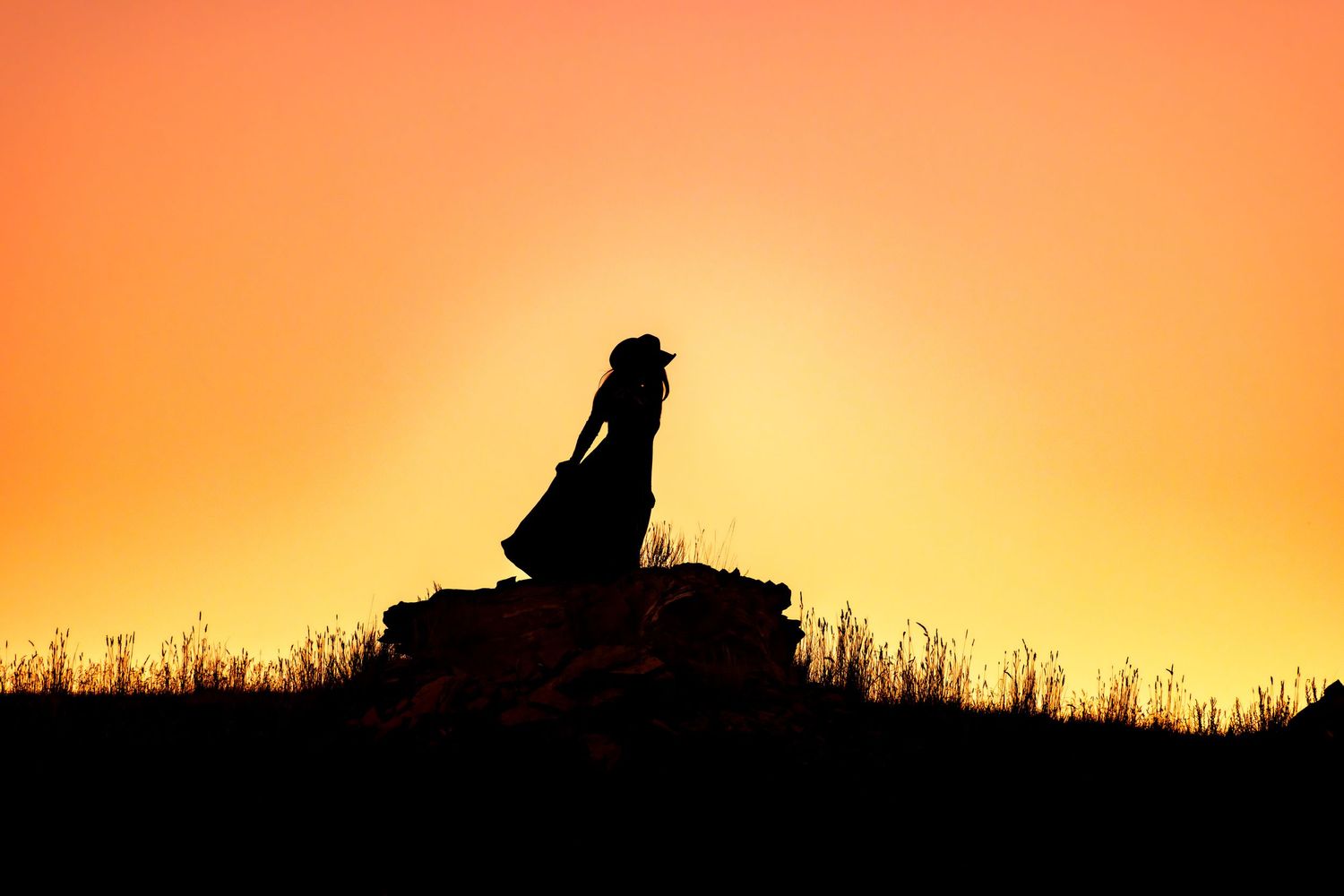Silhouette of a woman in a dress and hat standing on rocks against a vibrant orange sunset sky.