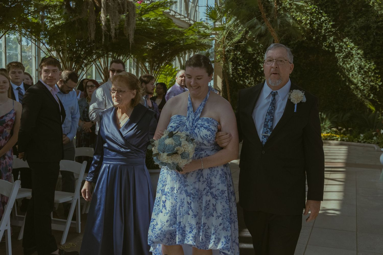 Bride in blue floral dress walks down the aisle with parents at an outdoor wedding ceremony.