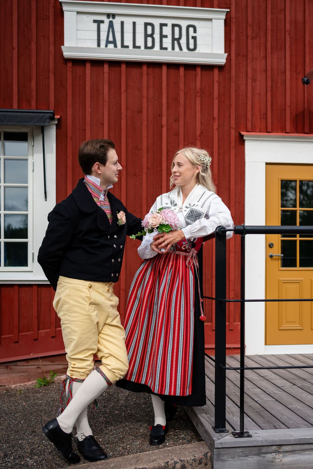 Couple in traditional Swedish folk costumes standing outside Tällberg building, woman holding pink flowers.