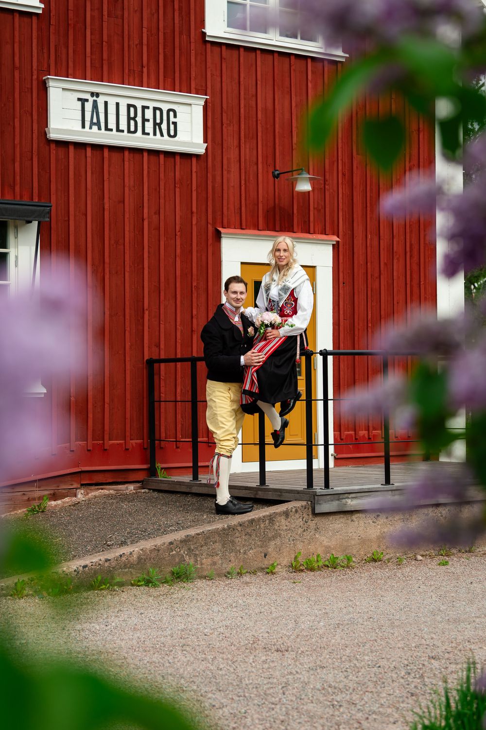 Couple posing on steps of red Tällberg building, woman in traditional Swedish costume holding flowers, framed by purple blooms.