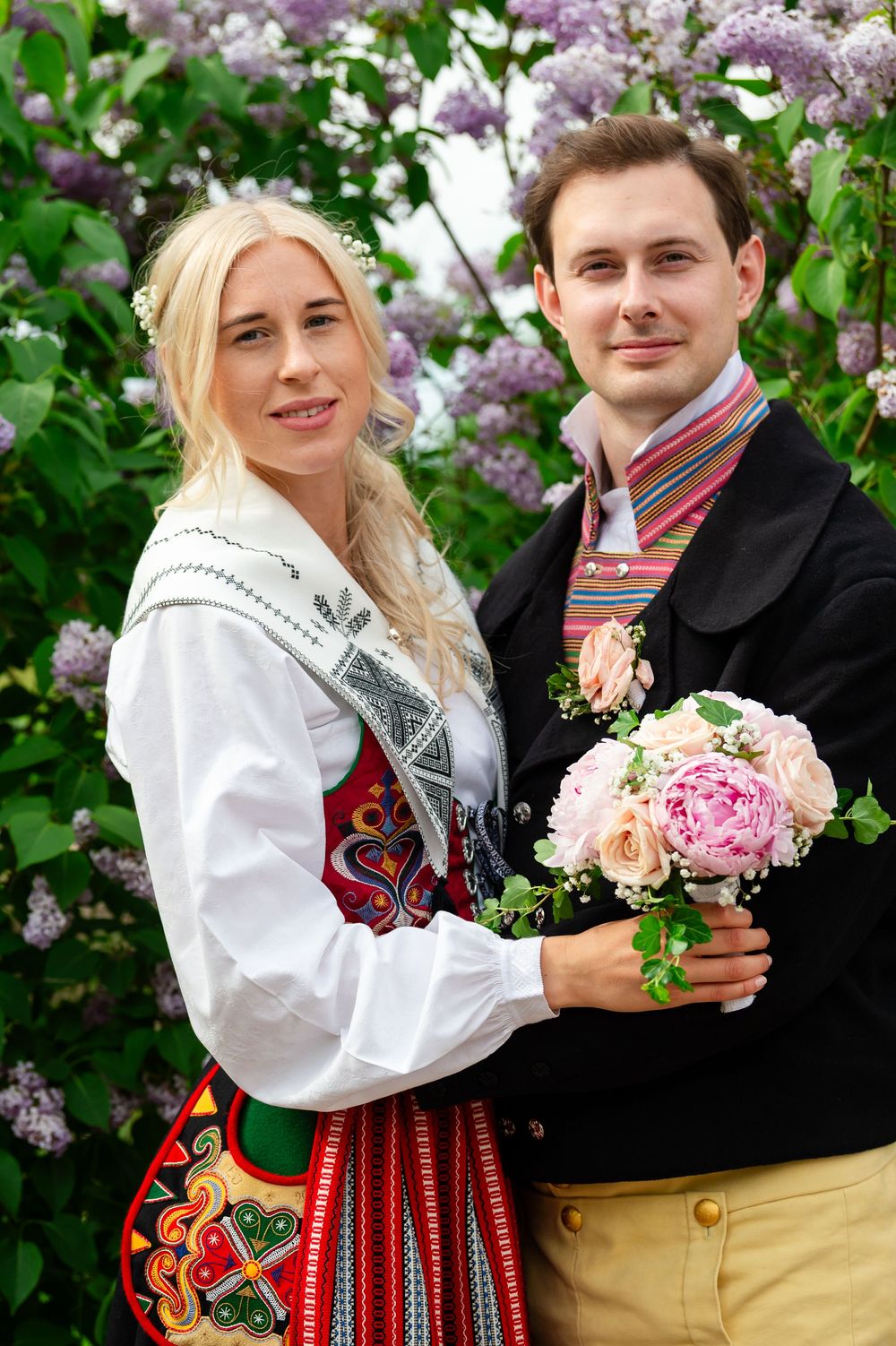 Couple in traditional Norwegian bunad attire posing with pink bouquet before blooming lilac bushes.