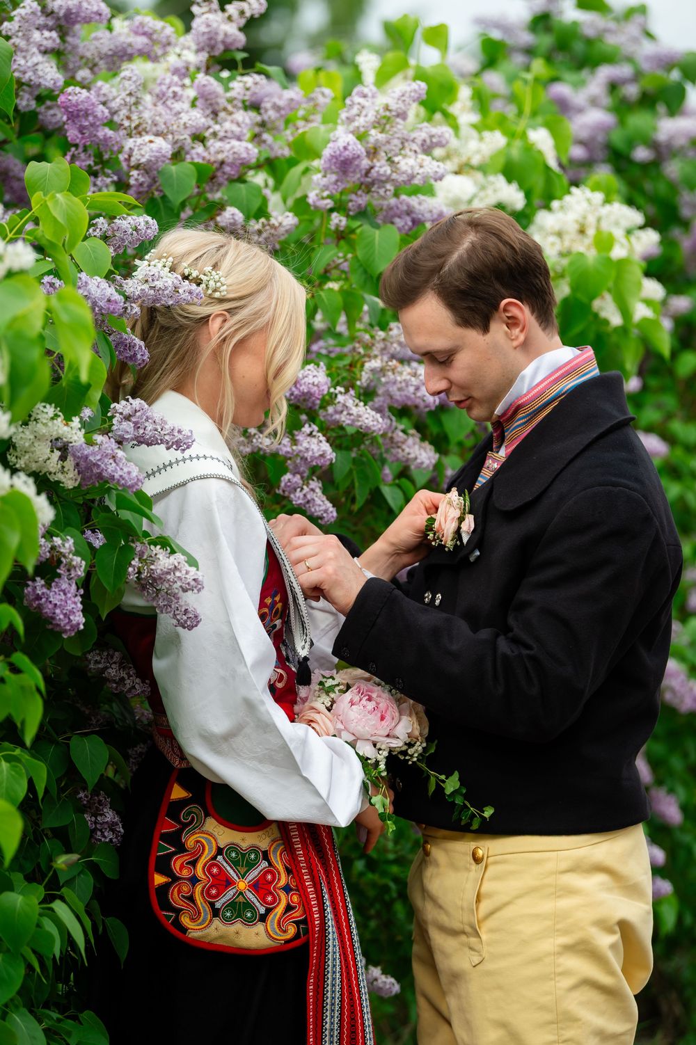 Couple in Norwegian traditional bunad attire standing among blooming lilac bushes.