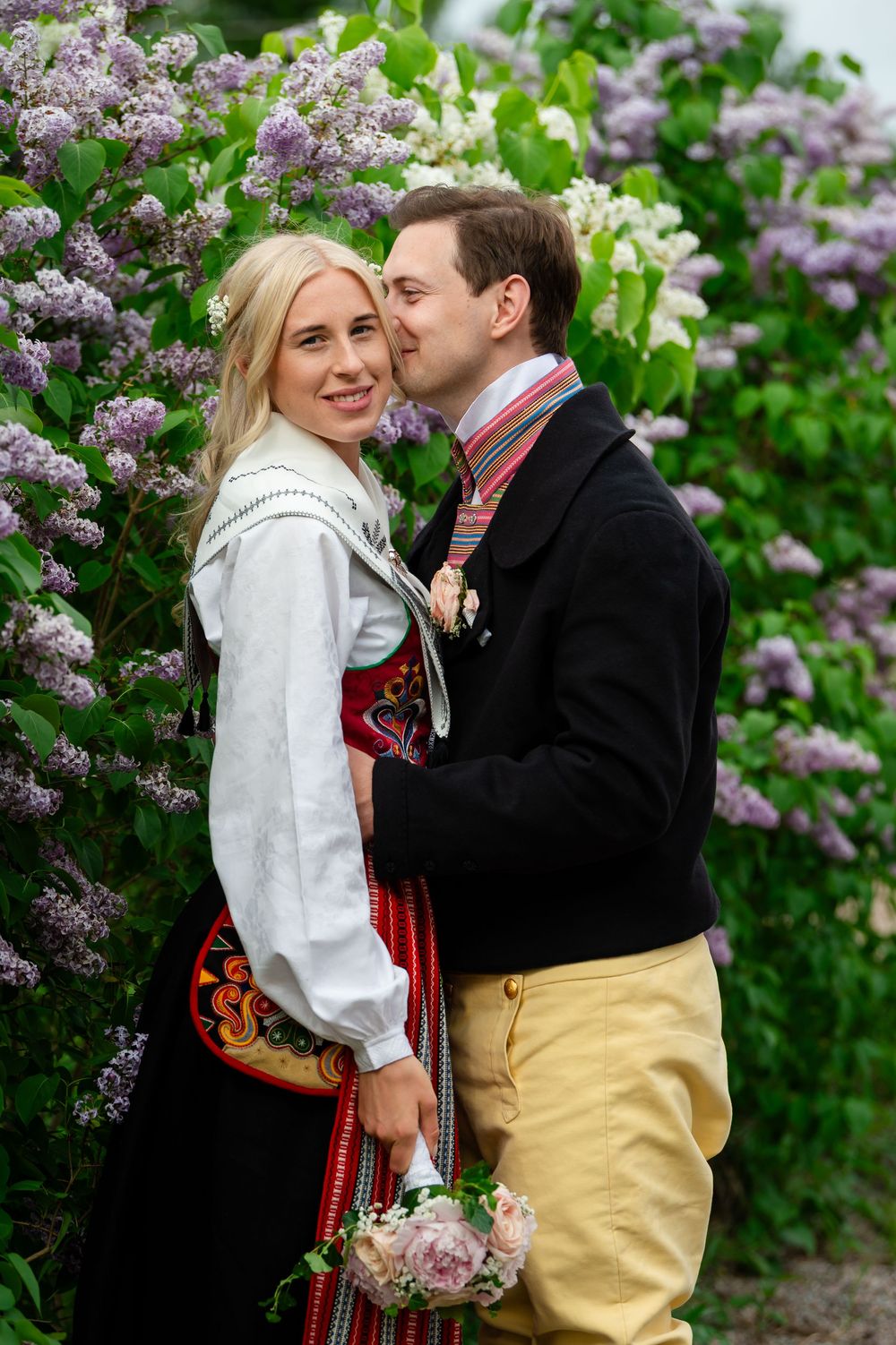Couple in Norwegian traditional bunad costumes embracing near blooming lilac bushes outdoors.
