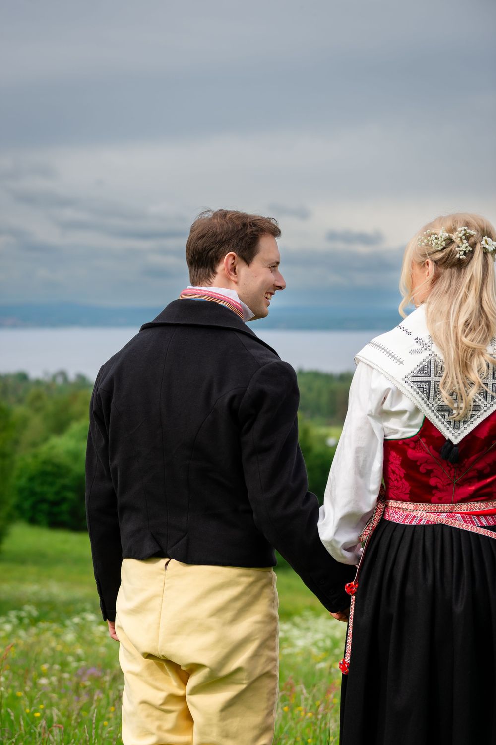 Couple in traditional Scandinavian folk costumes holding hands in a green meadow with cloudy sky backdrop.