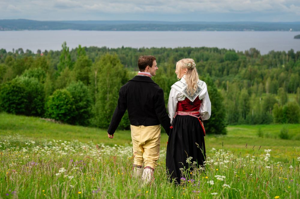 Couple in traditional Scandinavian folk costumes walking hand in hand through a green meadow with a lake view.