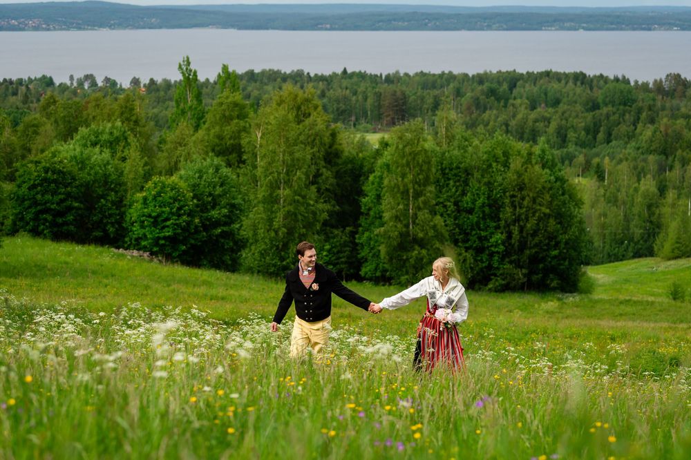 Two people in traditional folk costumes hold hands while walking through a lush green meadow with wildflowers and forest backdrop.