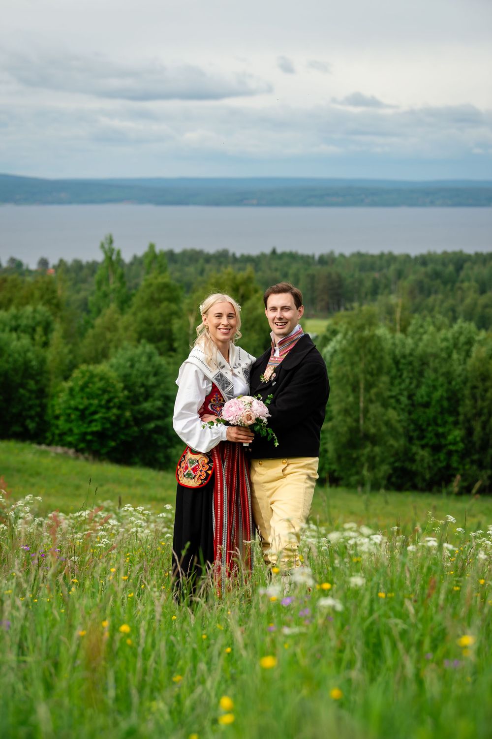 Couple in traditional Scandinavian folk costumes posing in a wildflower meadow with forest and lake views.