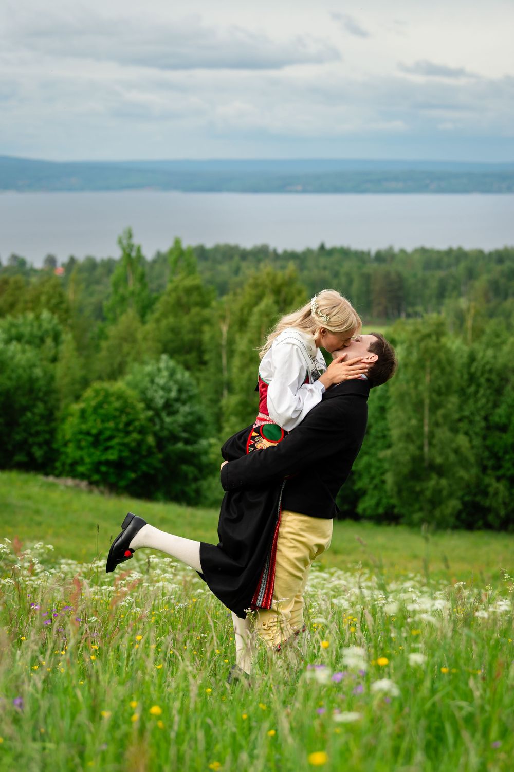 Man lifting woman in traditional Nordic attire for a romantic kiss in a scenic wildflower meadow.