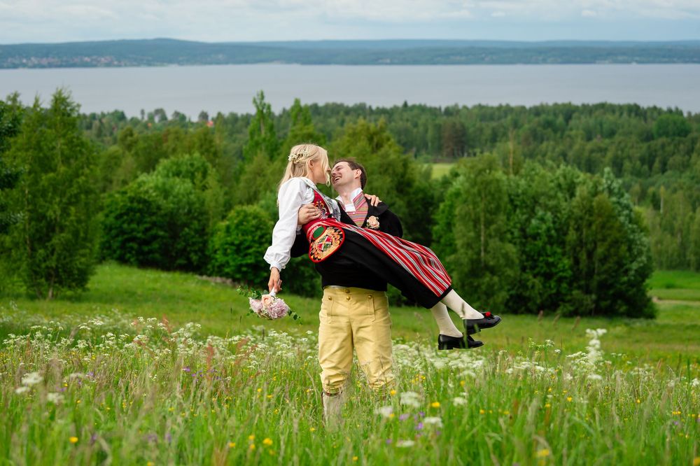 Couple in traditional Scandinavian folk costumes embracing in a green meadow with a lake and forest in the background.