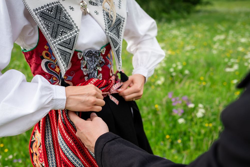 Person wearing traditional Nordic folk costume being helped to fasten a decorative red and black bunad outdoors.