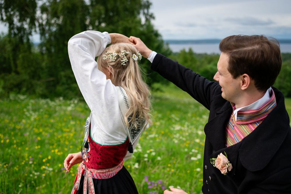 Man in traditional Norwegian bunad adjusting woman's floral hair wreath in green meadow.