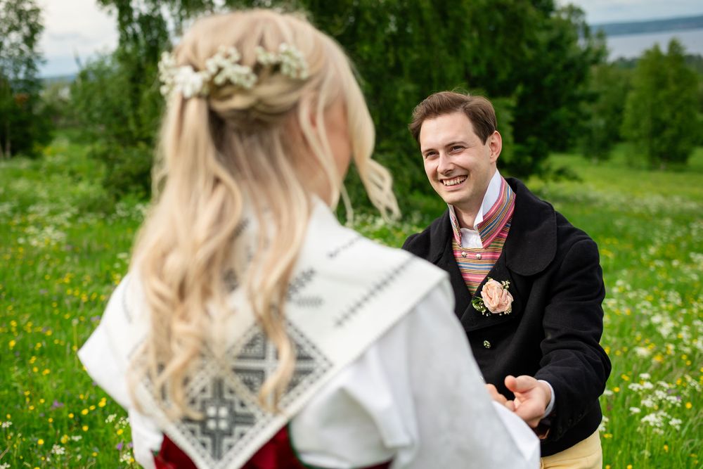 Smiling man in traditional Norwegian bunad costume holding hands with blonde woman in floral meadow.