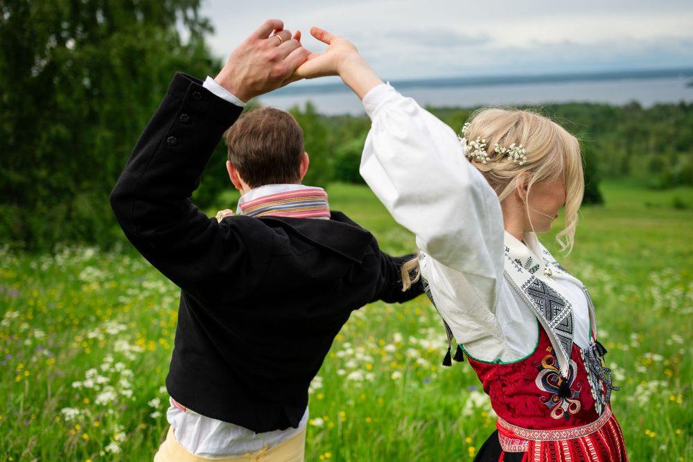 Couple in traditional Scandinavian folk costumes dancing outdoors in a green meadow with wildflowers and lake in background.