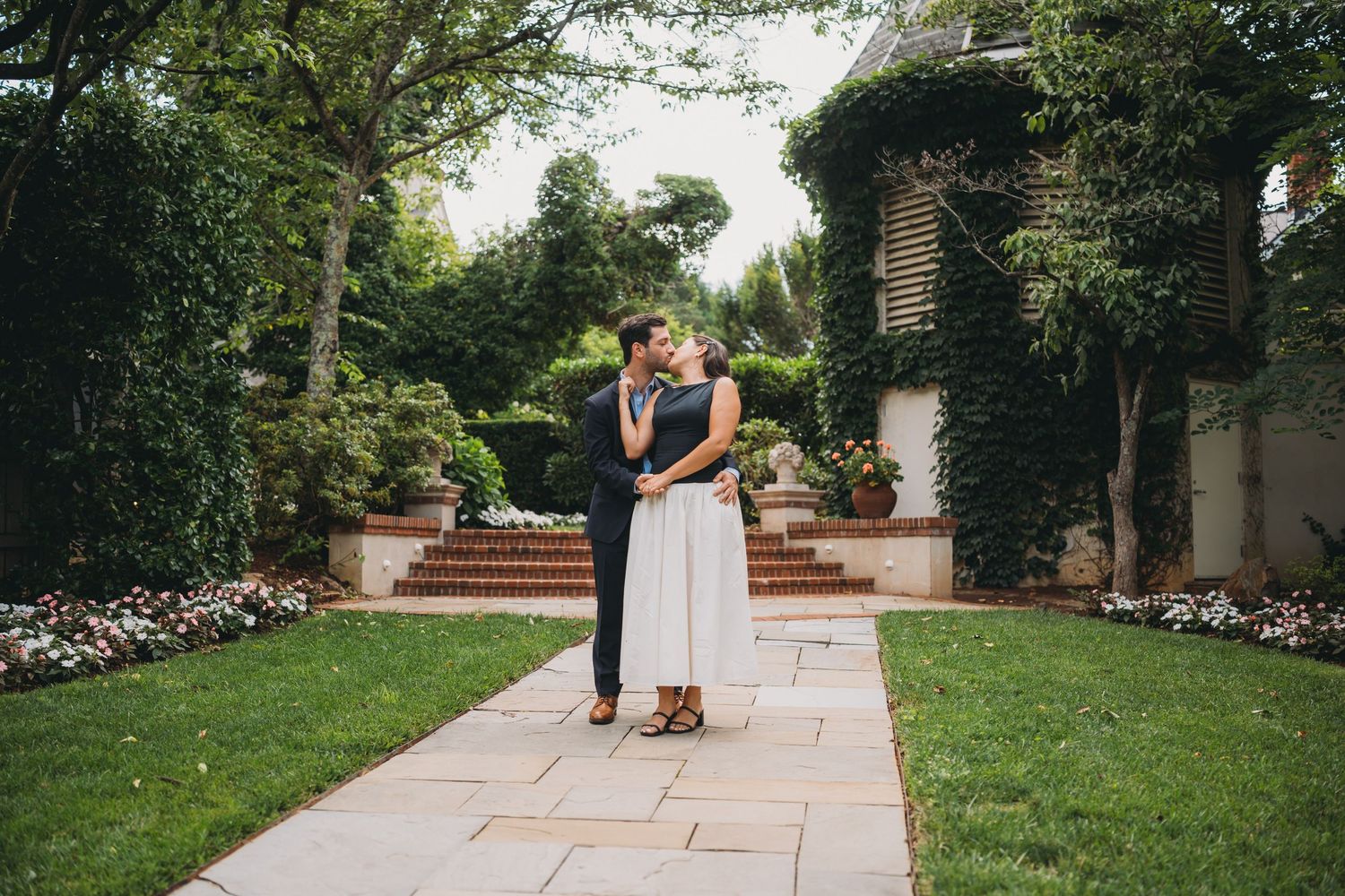 Couple holding hands walking through a lush garden with ivy-covered walls and brick steps.