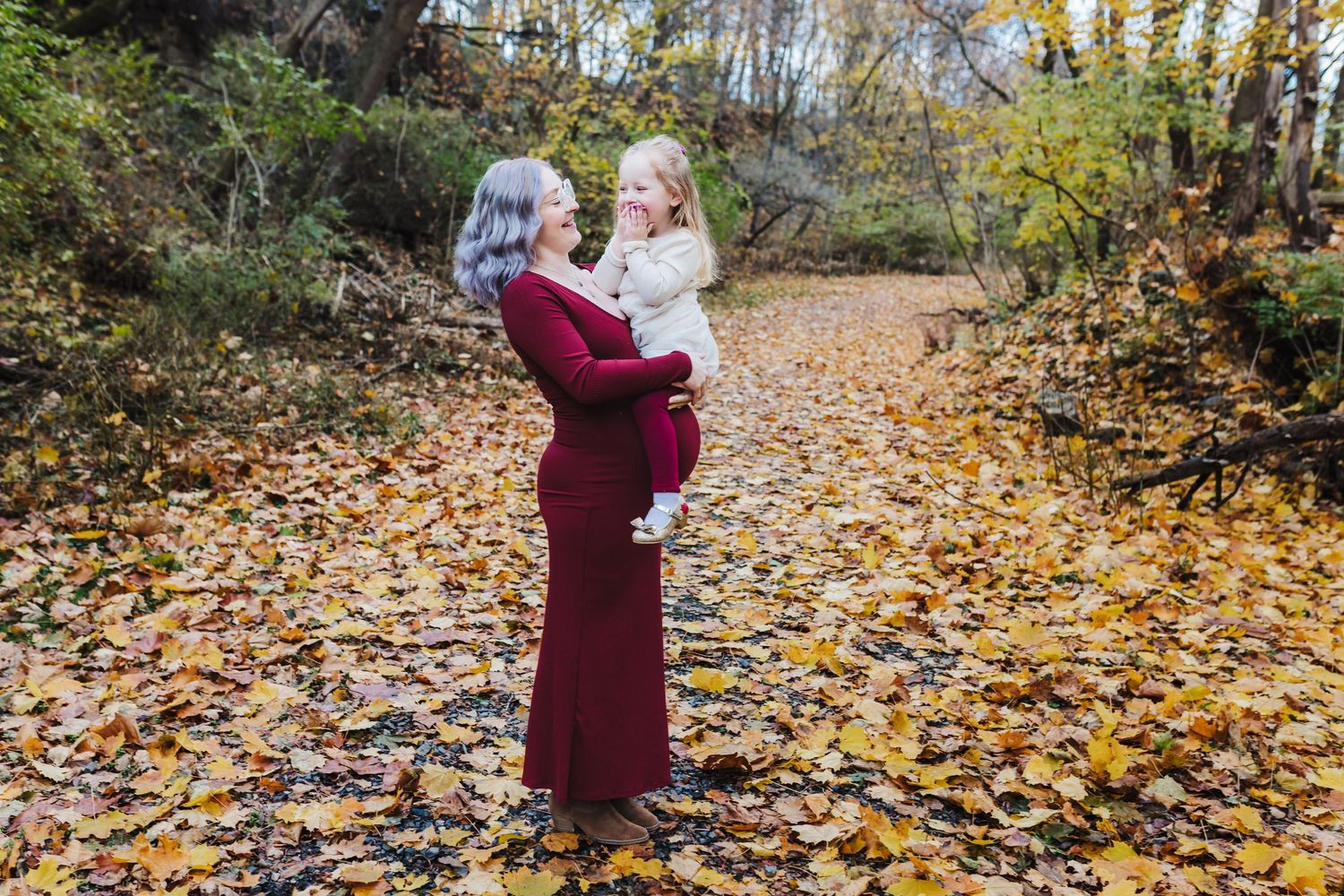 Pregnant woman in red gown holds toddler outdoors among golden autumn leaves on a forest path.