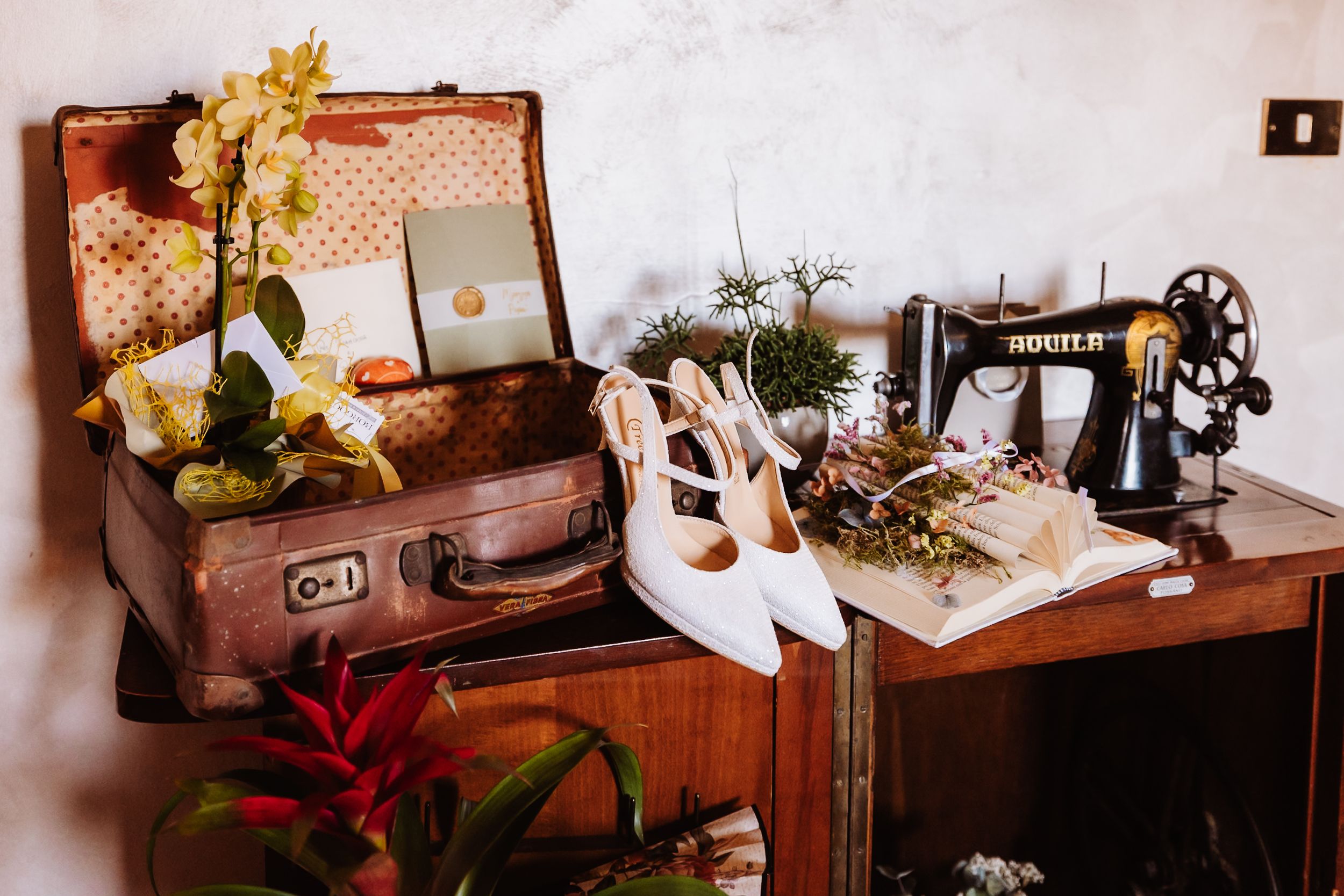 Vintage suitcase with orchids, white bridal heels, and antique sewing machine styled in a rustic wedding flat lay.