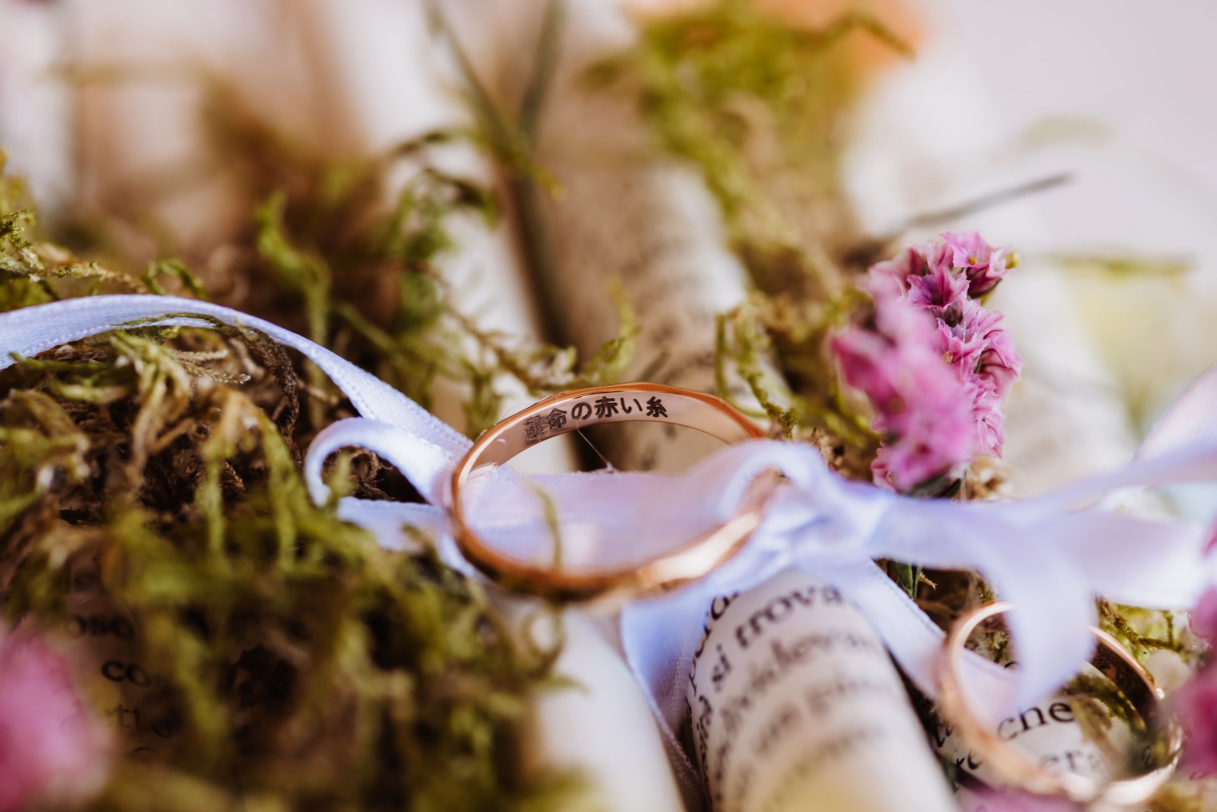 Gold wedding rings resting on a floral arrangement with moss, pink flowers, and ribbon accents.