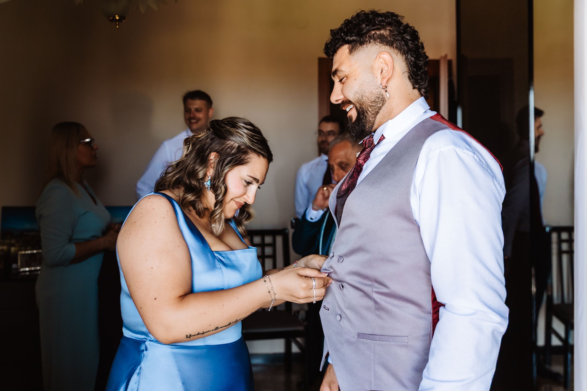 Woman in blue dress adjusting boutonniere on smiling groom in gray vest at wedding celebration.