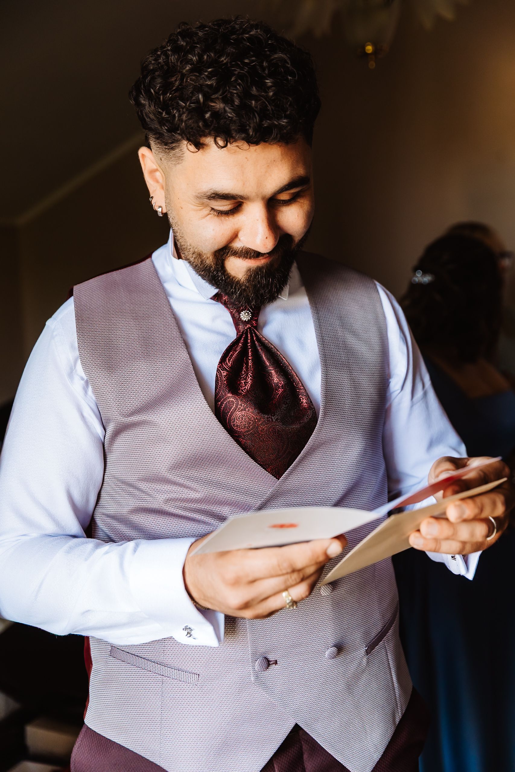 A bearded groom in a gray vest and burgundy cravat reads a heartfelt letter while smiling on his wedding day.